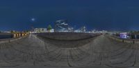 a fisheye photograph of a walkway and a body of water at night with a few buildings in the background