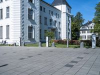 a paved street outside of some very big buildings and trees behind it and a white building with two stories behind the building