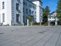 a paved street outside of some very big buildings and trees behind it and a white building with two stories behind the building