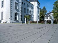 a paved street outside of some very big buildings and trees behind it and a white building with two stories behind the building