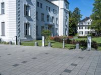 a paved street outside of some very big buildings and trees behind it and a white building with two stories behind the building