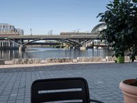 two chairs sitting next to a table with plants on a sidewalk near water and buildings