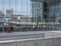 people walking past a train station in an open city square with tall buildings reflected in the windows