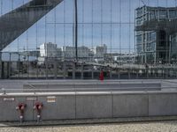 people walking past a train station in an open city square with tall buildings reflected in the windows