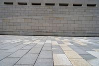 a brick wall next to an empty sidewalk in the afternoon suntime with sunlight reflecting off of windows and squares of tiles