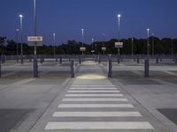 empty parking lot with street lights and poles at dusk, in background are trees and a blue sky