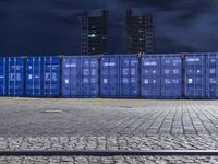 a group of blue shipping containers stacked against each other on a brick walkway at night