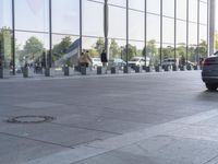 a black car driving past a glass building with several people walking on it, near parked cars and a walkway