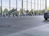 a black car driving past a glass building with several people walking on it, near parked cars and a walkway