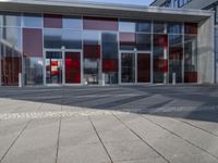 empty sidewalk outside a modern glass and concrete building with red accents for doors, windows, and street lights