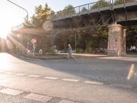 the intersection of two street roads and two bridges in a small city park with graffiti covered pillars, people on benches, parked cars and a pedestrian crossing over a roadway