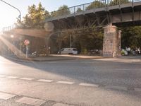 the intersection of two street roads and two bridges in a small city park with graffiti covered pillars, people on benches, parked cars and a pedestrian crossing over a roadway