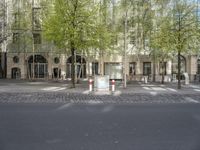 the side of a building, near a street lined with trees and chairs, has white boxes in front