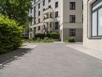 a brick walkway between two beige apartment buildings next to a tree filled street and some bushes