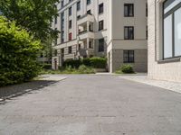 a brick walkway between two beige apartment buildings next to a tree filled street and some bushes