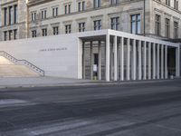 the large, empty sidewalk with multiple columns in front of the building with two signs