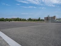 an empty parking lot with trees near by and the sky above it the pavement is smooth with white clouds in the distance