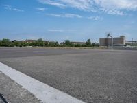 an empty parking lot with trees near by and the sky above it the pavement is smooth with white clouds in the distance