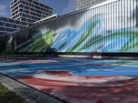 a group of people walk by some colorful art on a wall near tall buildings with metal poles