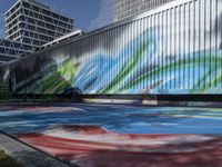 a group of people walk by some colorful art on a wall near tall buildings with metal poles