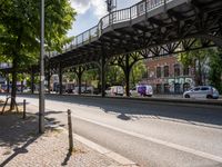 this is a city street under a bridge on a sunny day with parked cars, and people walking along