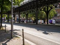 this is a city street under a bridge on a sunny day with parked cars, and people walking along