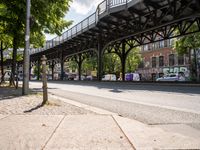 this is a city street under a bridge on a sunny day with parked cars, and people walking along