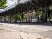 this is a city street under a bridge on a sunny day with parked cars, and people walking along