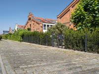 a paved cobbled street leads to several red brick buildings on either side of it and a fence with bushes lining both sides