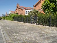 a paved cobbled street leads to several red brick buildings on either side of it and a fence with bushes lining both sides