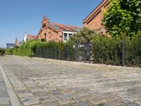 a paved cobbled street leads to several red brick buildings on either side of it and a fence with bushes lining both sides