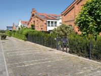 a paved cobbled street leads to several red brick buildings on either side of it and a fence with bushes lining both sides