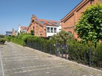 a paved cobbled street leads to several red brick buildings on either side of it and a fence with bushes lining both sides