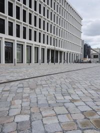 two people riding bikes on a city brick street in front of an empty building with white windows