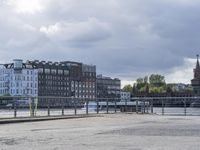 several benches on an asphalt surface next to a body of water under cloudy skies above the city