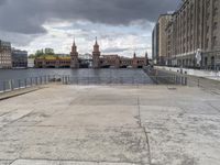 the man is standing on the sidewalk looking out at a canal in the city while a dark cloud is gathering over