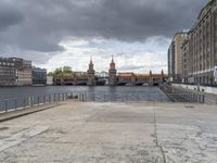 the man is standing on the sidewalk looking out at a canal in the city while a dark cloud is gathering over