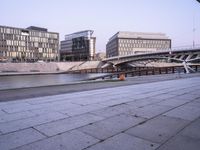 a stone walkway across from some buildings in a city with a bridge crossing over water