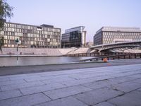 a stone walkway across from some buildings in a city with a bridge crossing over water