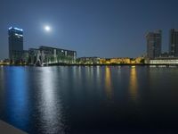 a night sky with buildings and a full moon in the sky over water in front of the building