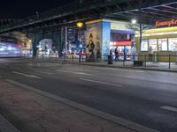 an intersection with many shops in the distance at night time, and a bus passing under an elevated overpass