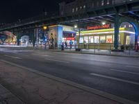 an intersection with many shops in the distance at night time, and a bus passing under an elevated overpass