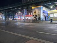 an intersection with many shops in the distance at night time, and a bus passing under an elevated overpass