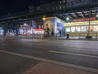 an intersection with many shops in the distance at night time, and a bus passing under an elevated overpass
