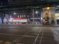 an intersection with many shops in the distance at night time, and a bus passing under an elevated overpass