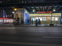 an intersection with many shops in the distance at night time, and a bus passing under an elevated overpass
