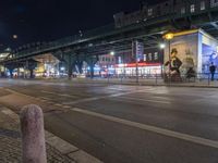 an intersection with many shops in the distance at night time, and a bus passing under an elevated overpass