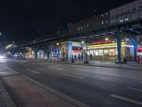 an intersection with many shops in the distance at night time, and a bus passing under an elevated overpass