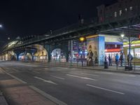 an intersection with many shops in the distance at night time, and a bus passing under an elevated overpass