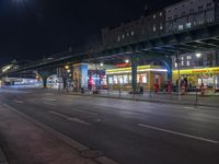 an intersection with many shops in the distance at night time, and a bus passing under an elevated overpass